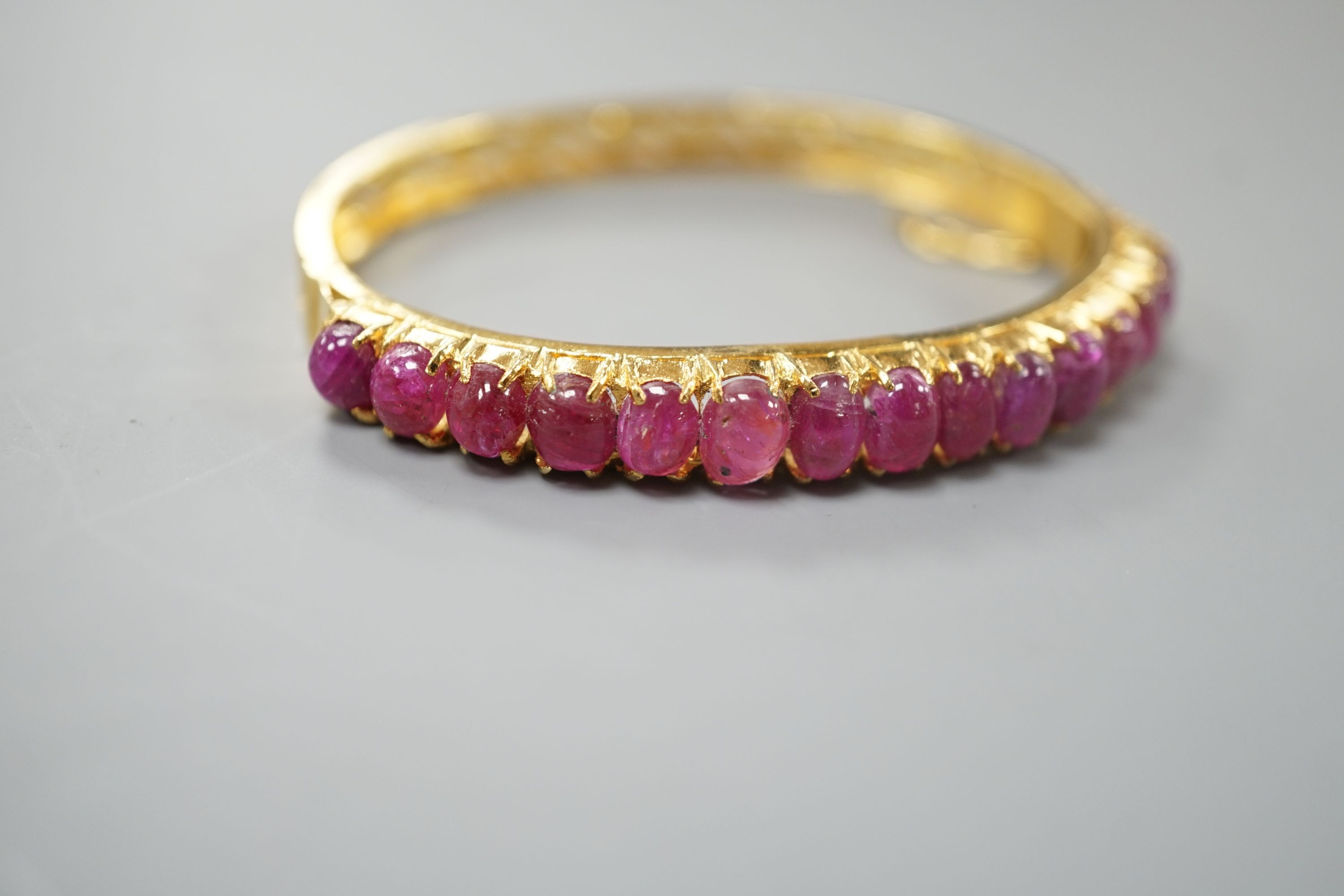 A modern Thai yellow metal and graduated fifteen stone cabochon ruby set hinged bangle, interior diameter 54mm, gross weight 12.9 grams.